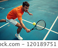 A focused male tennis player in an orange shirt is lunging to hit a yellow tennis ball with his racket on a blue court. The image captures the intensity of the game, showcasing the player's 120189751