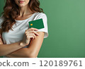 A young woman with wavy hair is holding a green credit card close to her chest against a solid green background. The image conveys a sense of financial empowerment and modern lifestyle, highlighting 120189761