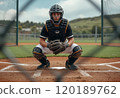A focused young male baseball catcher is crouched behind home plate, wearing a black helmet and protective gear. The image captures the intensity of the moment, framed by the diamond mesh of the fence 120189762