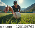 A focused baseball player in a black jersey and cap is reaching out towards a baseball on the grass field under a bright sun. The image captures the intensity of the moment, highlighting the player's 120189763