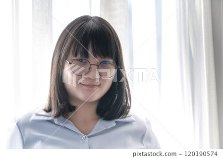 Asian glasses cute woman in blue shirt beside the curtain and window light in bedroom. 120190574