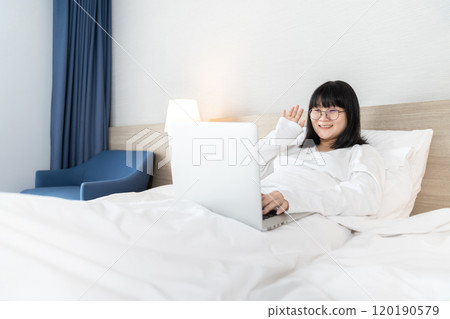 Glasses Asian young pretty teenager rise hand up hi and hello to laptop notebook for meeting and video conference on the bed in Work From Home period. 120190579