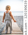 Faceless blond toddler in grey overalls stands alone on wooden path leads to seashore on summer day. 120190639