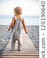 Beautiful toddler in summer grey jumpsuit stands alone on path leading to the seashore on warm day. 120190640