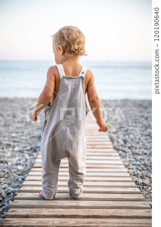 Beautiful toddler in summer grey jumpsuit stands alone on path leading to the seashore on warm day. 120190640
