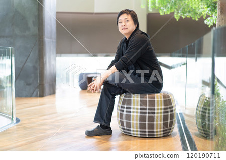 In the contemporary hall of a modern building, an Asian man dressed in black attire finds comfort as he sits on a circular sofa. In the contemporary hall of a modern building, an Asian man dressed in black attire finds comfort as he sits on a circular sofa. 120190711