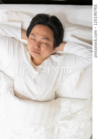 Enjoying a restful sleep, an Asian man finds comfort in his white long-sleeved cotton pajama shirt as he lies on a soft bed, nestled under a gentle blanket and supported by a plush pillow. Enjoying a restful sleep, an Asian man finds comfort in his white long-sleeved cotton pajama shirt as he lies on a soft bed, nestled under a gentle blanket and supported by a plush pillow. 120190768