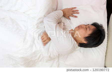 Enjoying a restful sleep, an Asian man finds comfort in his white long-sleeved cotton pajama shirt as he lies on a soft bed, nestled under a gentle blanket and supported by a plush pillow. Enjoying a restful sleep, an Asian man finds comfort in his white long-sleeved cotton pajama shirt as he lies on a soft bed, nestled under a gentle blanket and supported by a plush pillow. 120190772