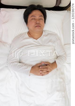 Enjoying a restful sleep, an Asian man finds comfort in his white long-sleeved cotton pajama shirt as he lies on a soft bed, nestled under a gentle blanket and supported by a plush pillow. Enjoying a restful sleep, an Asian man finds comfort in his white long-sleeved cotton pajama shirt as he lies on a soft bed, nestled under a gentle blanket and supported by a plush pillow. 120190774
