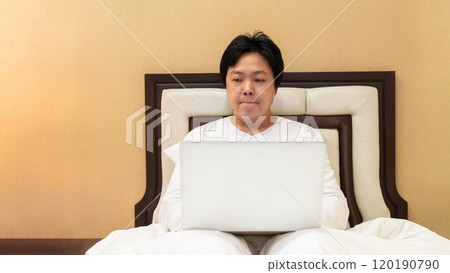 Work from home, a freelancer man leaning against the bed and using his laptop to work online on a bed. Work from home, a freelancer man leaning against the bed and using his laptop to work online on a bed. 120190790