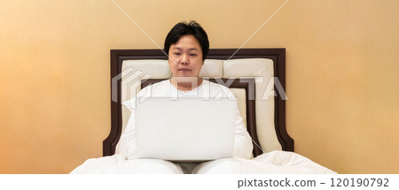Work from home, a freelancer man leaning against the bed and using his laptop to work online on a bed. Work from home, a freelancer man leaning against the bed and using his laptop to work online on a bed. 120190792