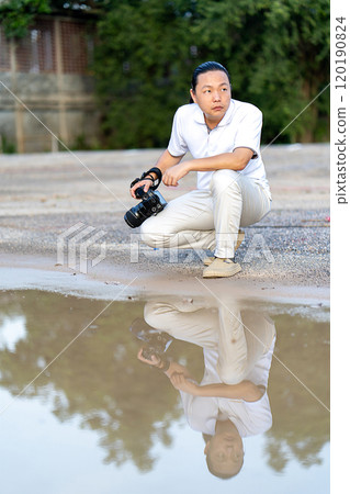 Asian man holds a big black mirrorless camera medium format type on cement asphalt ground in front of water reflex. 120190824