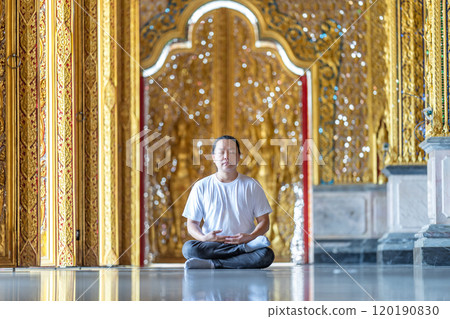 Asian Man meditates on the ground with reflection in the sunlight pouring into the temple church with gold gold Thai pattern blur background. 120190830