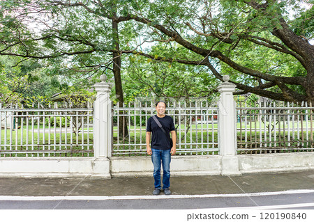 Asian Black T-Shirt is standing in front of white vintage fence beside the road in the afternoon time. 120190840