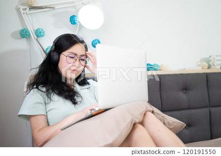 Glasses beautiful Asian woman is working on her laptop in the bedroom with a pastel green - brown color theme. Glasses beautiful Asian woman is working on her laptop in the bedroom with a pastel green - brown color theme. 120191550