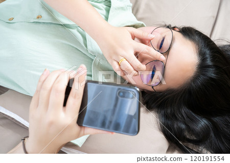Beautiful glasses Asian woman lie on her back, chatting, typing, and texting, mobile in her bedroom with a pastel green - brown color theme. 120191554