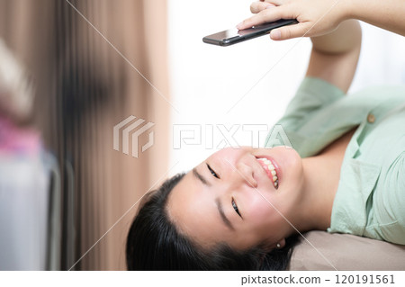Beautiful Asian woman is lying down and playing mobile on the bed in her bedroom with a pastel green - brown color theme. 120191561