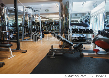 A well lit gym interior features weight machines, free weights, treadmills, and mirrors. Natural light streams through large windows onto a polished floor. A well lit gym interior features weight machines, free weights, treadmills, and mirrors. Natural light streams through large windows onto a polished floor. 120195651