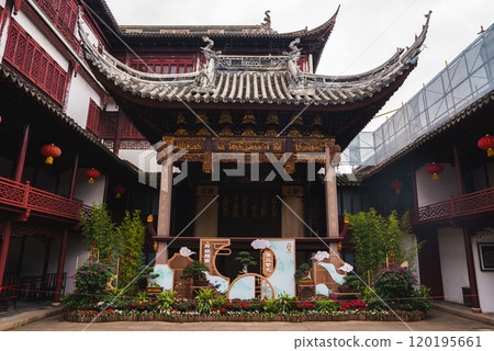 A traditional Chinese building in Shanghai features ornate carvings and upturned eaves. The courtyard is decorated with red lanterns and vibrant plants. 120195661