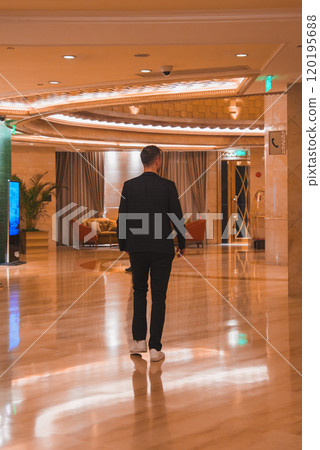 A man in a dark suit walks through a luxurious lobby with marble floors, modern decor, and intricate lighting fixtures in Shanghai. A man in a dark suit walks through a luxurious lobby with marble floors, modern decor, and intricate lighting fixtures in Shanghai. 120195688