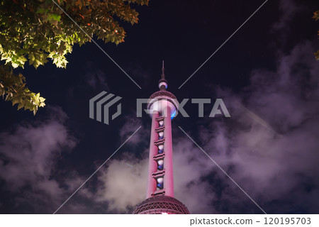 The Oriental Pearl Tower stands illuminated against a cloudy night sky in Shanghai, China, with tree leaves framing the scene in the foreground. The Oriental Pearl Tower stands illuminated against a cloudy night sky in Shanghai, China, with tree leaves framing the scene in the foreground. 120195703