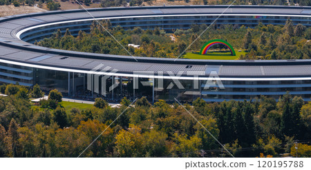 The image captures an aerial view of Apple Park in Cupertino, California, featuring the circular building, lush greenery, and a colorful rainbow arch. 120195788