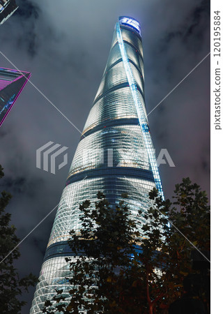 The Shanghai Tower stands illuminated against a cloudy night sky, showcasing its spiraling design and glass facade. Trees in the foreground add a natural touch. The Shanghai Tower stands illuminated against a cloudy night sky, showcasing its spiraling design and glass facade. Trees in the foreground add a natural touch. 120195884