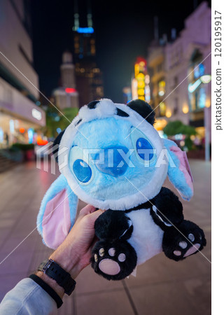 A blue plush toy with large eyes is held in the foreground, set against the illuminated skyscrapers and vibrant streets of Shanghai at night. 120195917