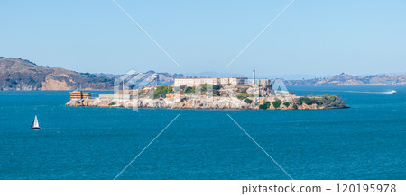 Alcatraz Island with its historic penitentiary and watchtower is surrounded by San Francisco Bay's blue waters. A sailboat and Marin Headlands are visible. 120195978