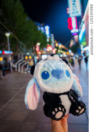 A bustling street in Shanghai at night features bright neon lights and busy pedestrians. A plush toy with blue eyes and floppy ears is held in the foreground. A bustling street in Shanghai at night features bright neon lights and busy pedestrians. A plush toy with blue eyes and floppy ears is held in the foreground. 120195999