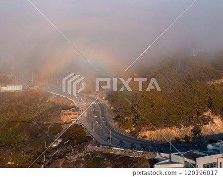 Aerial view of a foggy landscape in San Francisco, featuring a winding road, trees, buildings, and a faint rainbow arc in the mist. Aerial view of a foggy landscape in San Francisco, featuring a winding road, trees, buildings, and a faint rainbow arc in the mist. 120196017
