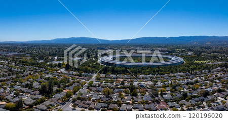 Aerial view of Apple Park in Cupertino, California, with its circular design, surrounded by greenery, residential neighborhoods, and distant mountains. 120196020