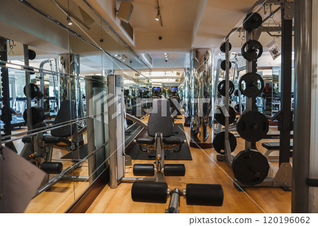 A well equipped gym interior featuring weight machines, free weights, mirrored walls, and wooden flooring, emphasizing strength training equipment. 120196062