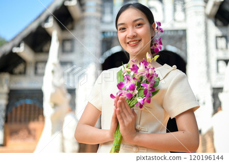 A beautiful, smiling Asian women stand in a temple, holding flowers in a prayer posture. 120196144