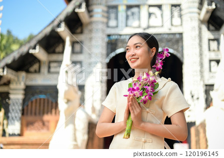 A smiling Asian women in traditional dresses stand in a temple, holding flowers in a prayer posture. 120196145
