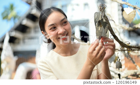 Smiling Asian woman hanging a good luck leaf on a tree in a temple, making a wish, visiting a temple 120196149