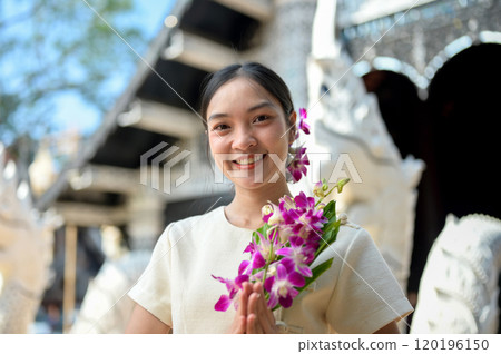 A beautiful, smiling Asian women stand in a temple, holding flowers in a prayer posture. 120196150