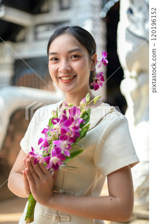 A beautiful, smiling Asian women stand in a temple, holding flowers in a prayer posture. 120196152