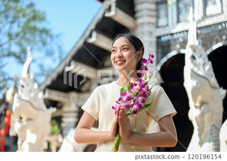 A smiling Asian women in traditional dresses stand in a temple, holding flowers in a prayer posture. A smiling Asian women in traditional dresses stand in a temple, holding flowers in a prayer posture. 120196154