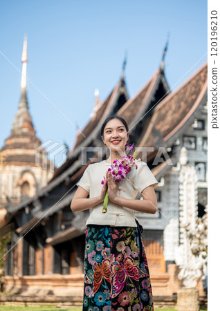 Charming Asian woman in a traditional dress stands outdoors with an ancient temple in the background Charming Asian woman in a traditional dress stands outdoors with an ancient temple in the background 120196210