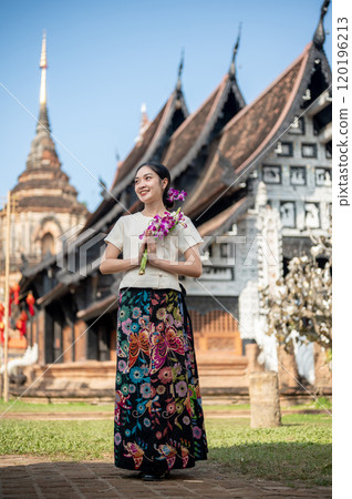 Charming Asian woman in a traditional dress stands outdoors with an ancient temple in the background 120196213