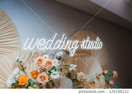A neon sign reading 'wedding studio' is surrounded by peach and white roses. The floral arrangements are set against pleated, fan like structures. 120196222