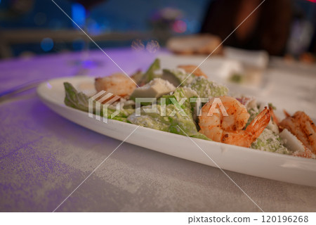A Caesar salad topped with grilled shrimp is served on a white plate. The background is softly blurred, indicating a dining setting with ambient lighting. 120196268