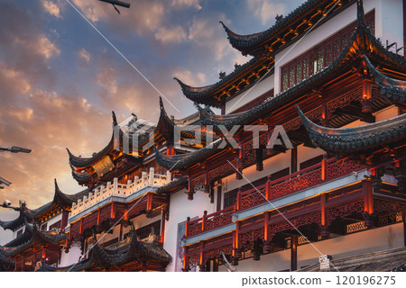 Intricately designed buildings with upturned eaves and ornate details are illuminated by warm lighting against a dramatic evening sky in Shanghai. 120196275