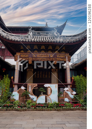 A traditional Chinese building in Shanghai features intricate wooden carvings and a curved roof with dragon motifs, surrounded by greenery and flowers. A traditional Chinese building in Shanghai features intricate wooden carvings and a curved roof with dragon motifs, surrounded by greenery and flowers. 120196288