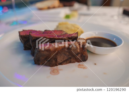 A plate displays slices of medium rare steak with a pink interior and seared exterior, accompanied by a small dish of dark sauce. Background is softly blurred. 120196290