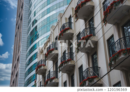 A sleek skyscraper with reflective glass stands beside a classic building with ornate balconies and red flowers, set against a clear blue sky in Shanghai. A sleek skyscraper with reflective glass stands beside a classic building with ornate balconies and red flowers, set against a clear blue sky in Shanghai. 120196369