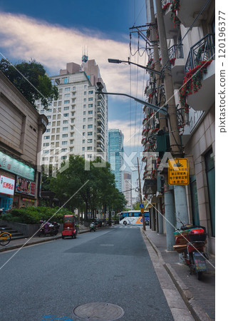 A quiet street in Shanghai, China, features buildings with flower adorned balconies, parked scooters, and a red tuk tuk, set against modern skyscrapers. 120196377