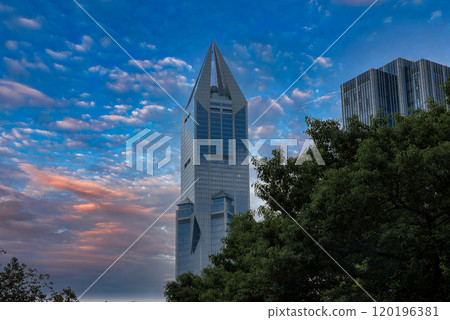A striking skyscraper with angular design stands against a vibrant sunset sky in Shanghai, partially obscured by lush green trees, with another high rise nearby. A striking skyscraper with angular design stands against a vibrant sunset sky in Shanghai, partially obscured by lush green trees, with another high rise nearby. 120196381