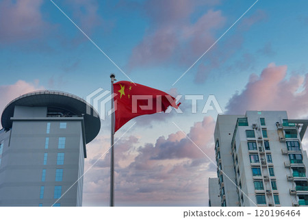 A Chinese flag waves prominently with a pink and blue sky backdrop. A modern building with a circular rooftop and a high rise residential building are visible. 120196464